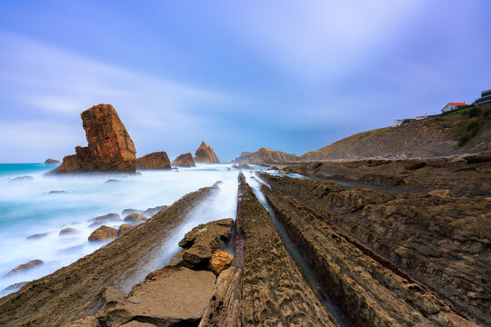 View of layered rock formations and sea stacks along the coast with long exposure waves crashing against the shore in Spain.