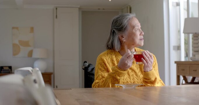 Senior Asian woman in wheelchair in yellow sweater stirring red cup sipping soon