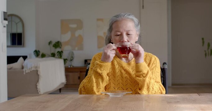 Senior Asian woman in wheelchair wearing mustard sweater lifting teacup to sip at home gazing right