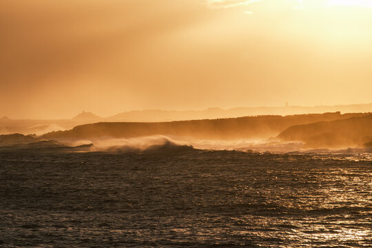 View of crashing ocean waves against a rocky coastline during a warm golden hour sunset in Spain.