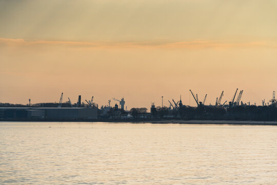 View of the industrial port with numerous cranes silhouetted against the soft golden light of sunset over the water in Gdansk, Pomeranian Voivodeship, Poland.