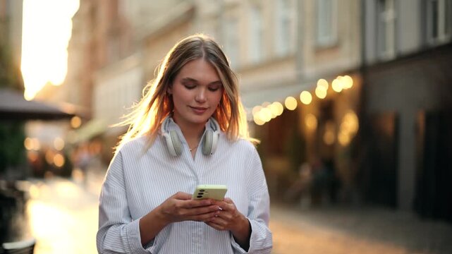 Beautiful attractive young blonde woman with headphones around neck walking on city street and texting on smartphone. Pretty female chatting online on blurred urban sunset background