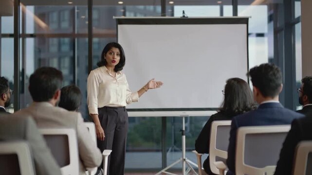 Starting meeting room woman pointing in silk top and pants explaining to audience with blank screen