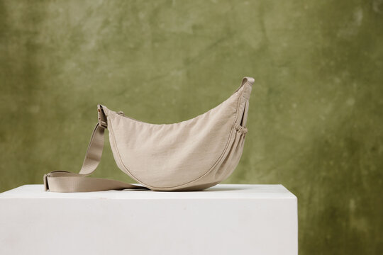 Stylish beige crescent shoulder bag with strap standing on white cube stand against textured olive green background studio product photography minimal modern design casual fabric handbag nylon purse a