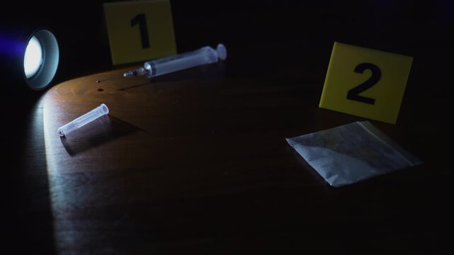 Drug baggie, syringe, and evidence markers scanned back and forth under flashlight and flashing police lights, point of view panning shot