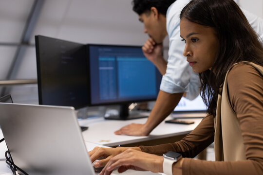 Diverse coworkers in business wear coding at desk, woman typing laptop while man viewing monitors