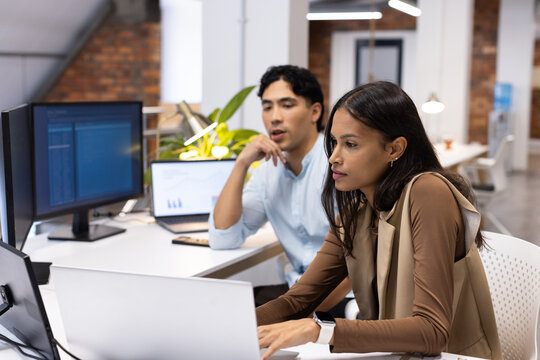 Diverse coworkers reviewing code, charts at desk, woman in brown top typing on laptop, copy space