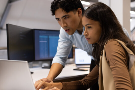 Diverse coworkers reviewing code on laptop at desk with dual monitors and phone, wearing shirt