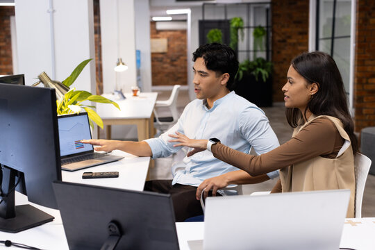 Diverse coworkers wearing light-blue shirt and beige vest analyzing laptop chart at open-plan desk