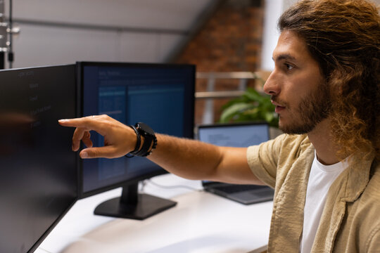 Man with curly hair pointing at monitor while using dual screens and laptop at modern desk
