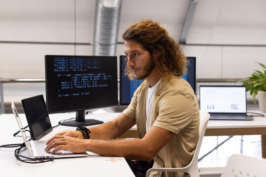 Adult man in beige overshirt, white tee typing on laptop at open-plan office with code monitor