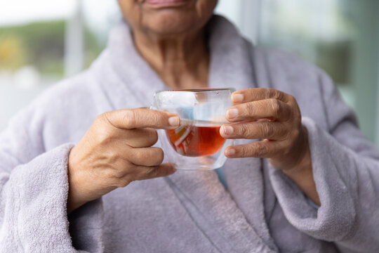 Senior African American woman holding double-walled glass cup of amber tea near window in bathrobe