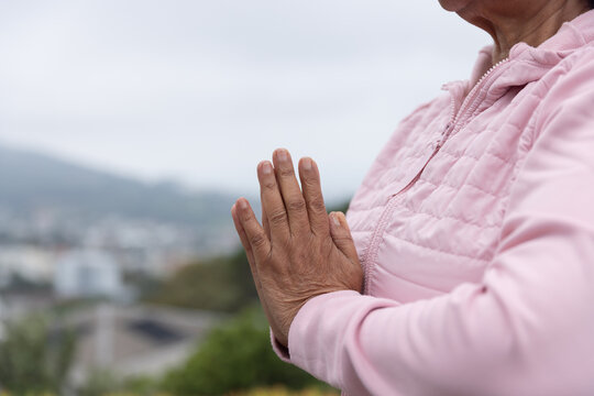 Senior woman pressing palms together at chest on hillside overlook wearing pink jacket, copy space