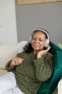Senior African American woman reclining on sofa at home in green sweater touching rose-pink headset