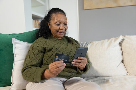 Senior African American woman wearing sweater checking phone holding credit card on couch at home