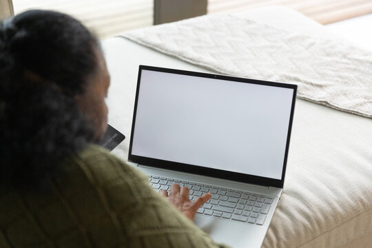 Senior African American woman in green sweater sitting on bed typing laptop holding card
