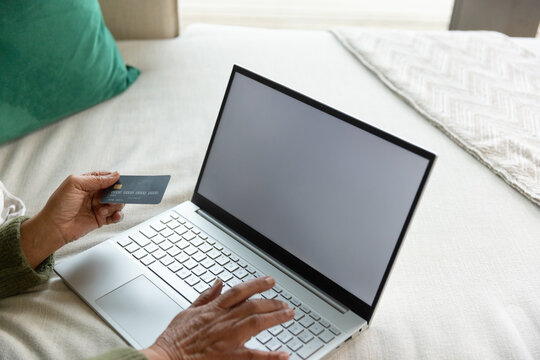 Senior African American woman holding credit card, typing on laptop in bed wearing green sweater