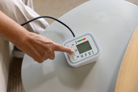 Senior African woman pressing start on BP-monitor, cuff tubing on side-table at home in light pants