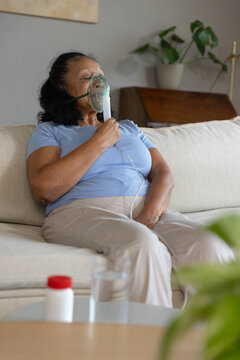 Senior African American woman sitting on couch wearing blue shirt holding mask and pill bottle