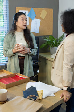 Diverse female coworkers in businesswear standing at office desk discussing fabric samples and mugs