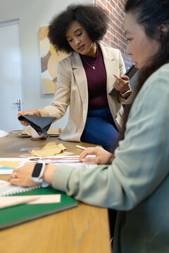 Diverse female coworkers in studio, one wearing beige blazer, examining black swatch and sketches