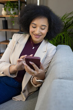 African American woman in blazer sitting on light-gray sofa holding maroon smartphone smiling