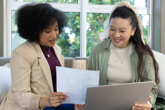 Diverse female friends sitting on sofa by window reviewing document, using laptop and holding mug