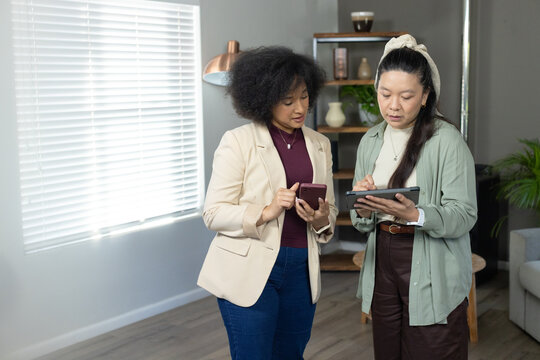 Diverse female coworkers in blazer reviewing on phone and tablet with stylus near window