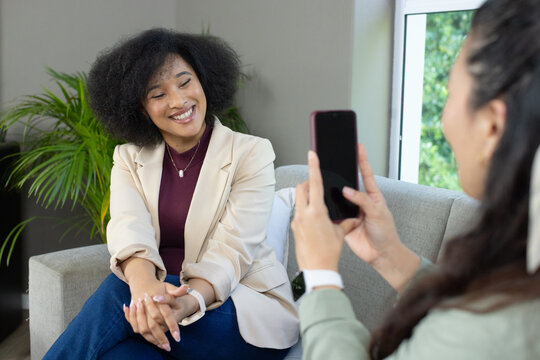 Diverse women sitting on sofa at home, one photographing friend wearing beige blazer with phone