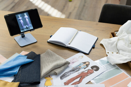 Wooden table is holding fabric swatches, sketches, notebook with pencil, tablet on stand in studio