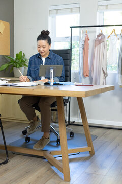Asian woman writing in notebook while referencing tablet on stand in home studio with clothing rack