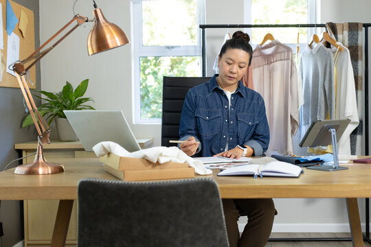 Asian woman sitting at wooden desk sketching fashion ideas in home studio with laptop