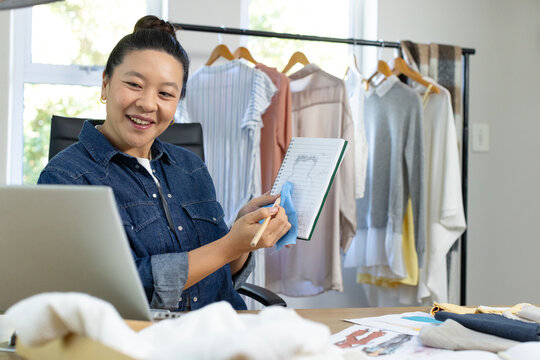 Asian woman in denim shirt holding sketchbook and blue swatch, sketching at desk near laptop
