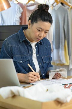 Asian woman wearing denim shirt sketching and inspecting fabric swatches at desk with laptop