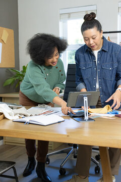 Diverse female coworkers examining fabric swatches and using tablet at bright design studio