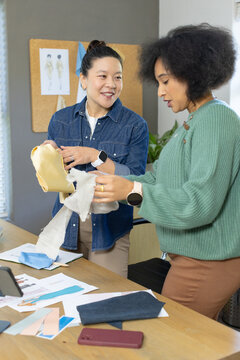 Diverse female designers examining fabric samples and sketches at wooden work table with smartphone