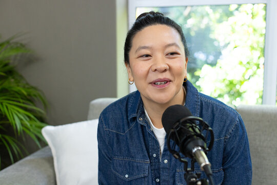 Woman speaking into microphone with pop filter on gray sofa in home studio, wearing denim shirt
