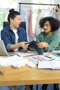 Asian and African American women examining black fabric sample with laptop and sketches at studio