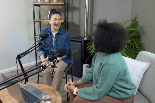 Diverse female co-hosts recording podcast in home studio at round table with mics and laptop