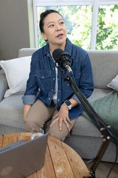 Asian woman speaking into microphone while sitting on sofa in living room with laptop