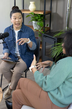 Diverse female hosts talking in home studio, denim shirt and green sweater, using tablet and mic