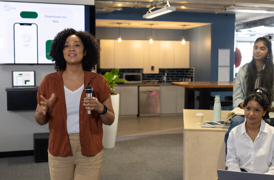 Female coworkers watching woman presenter wearing rust top holding microphone by display at kitchen