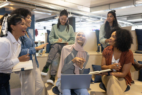 Diverse female team talking with woman wearing pink headscarf, using laptop and notebook in office