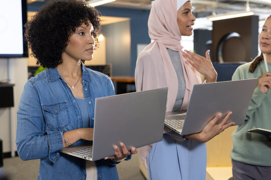African American coworkers meeting in office wearing denim shirt, headscarf holding silver laptops