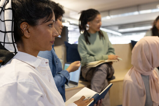 Diverse coworkers taking notes with laptop in lounge, woman in white top print headscarf