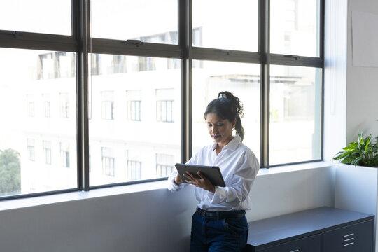 Female worker leaning on sill holding tablet by windows wearing white shirt dark pants, copy space