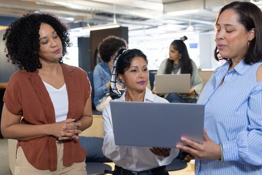 Female team gathering around presenter holding laptop, discussing in office in business wear