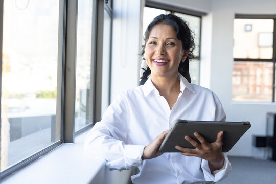 Indian woman holding tablet smiling by windowsill in office wearing white shirt and patterned scarf