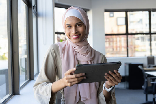 Woman in twenties standing by large windows in office holding dark tablet wearing pale pink hijab