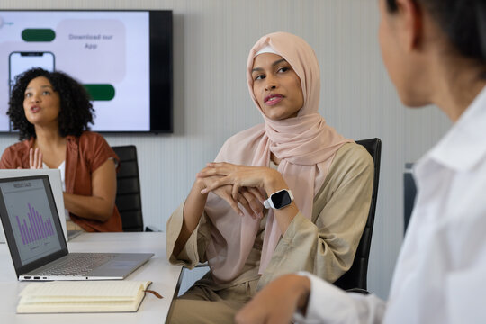 Diverse female coworkers discussing slides at meeting table with laptop, pink hijab, smartwatch